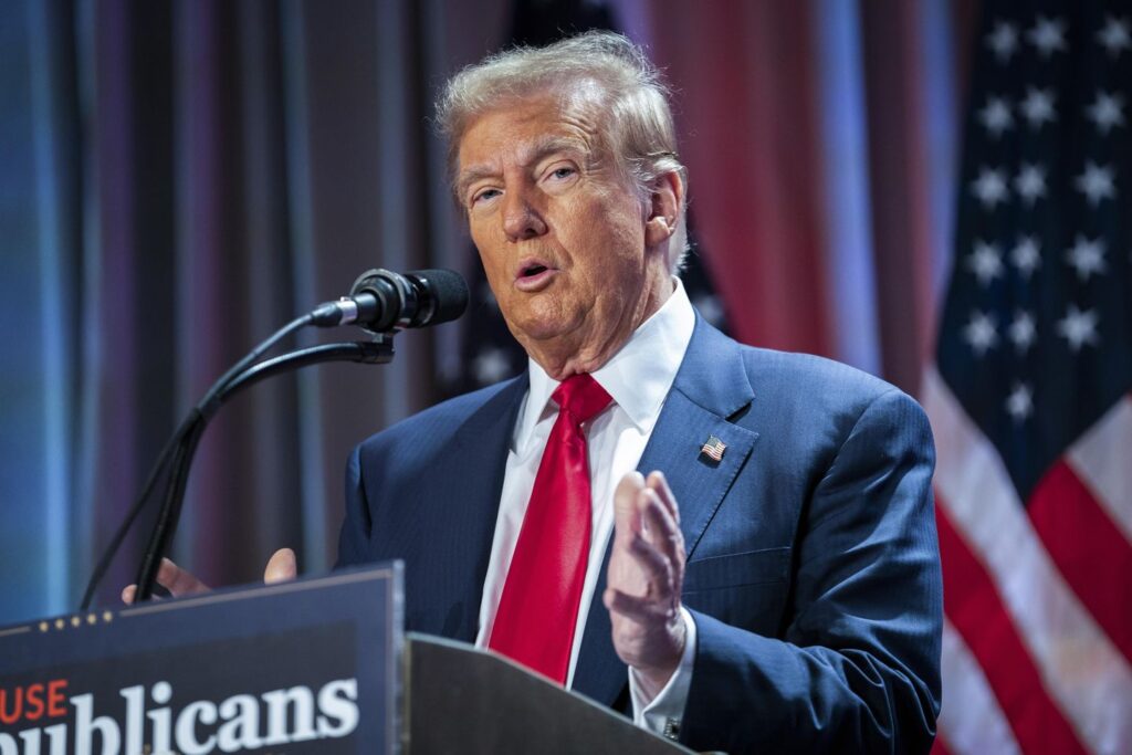 A man in a suit and red tie is speaking at a podium.