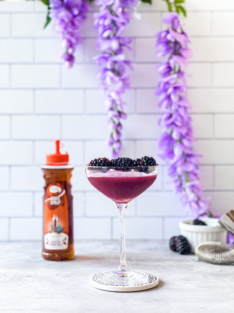 A purple drink with blackberries in it next to a bottle of syrup.