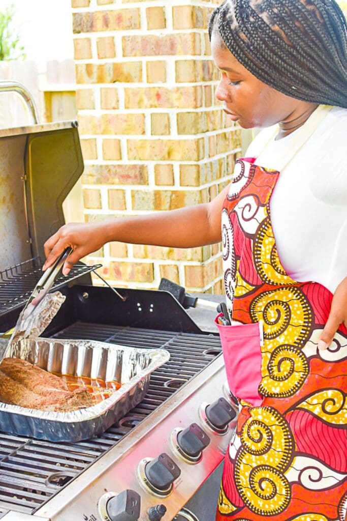Jazzmine tending to rack of baby back ribs on a gas grill while wearing a colorful ankara apron.