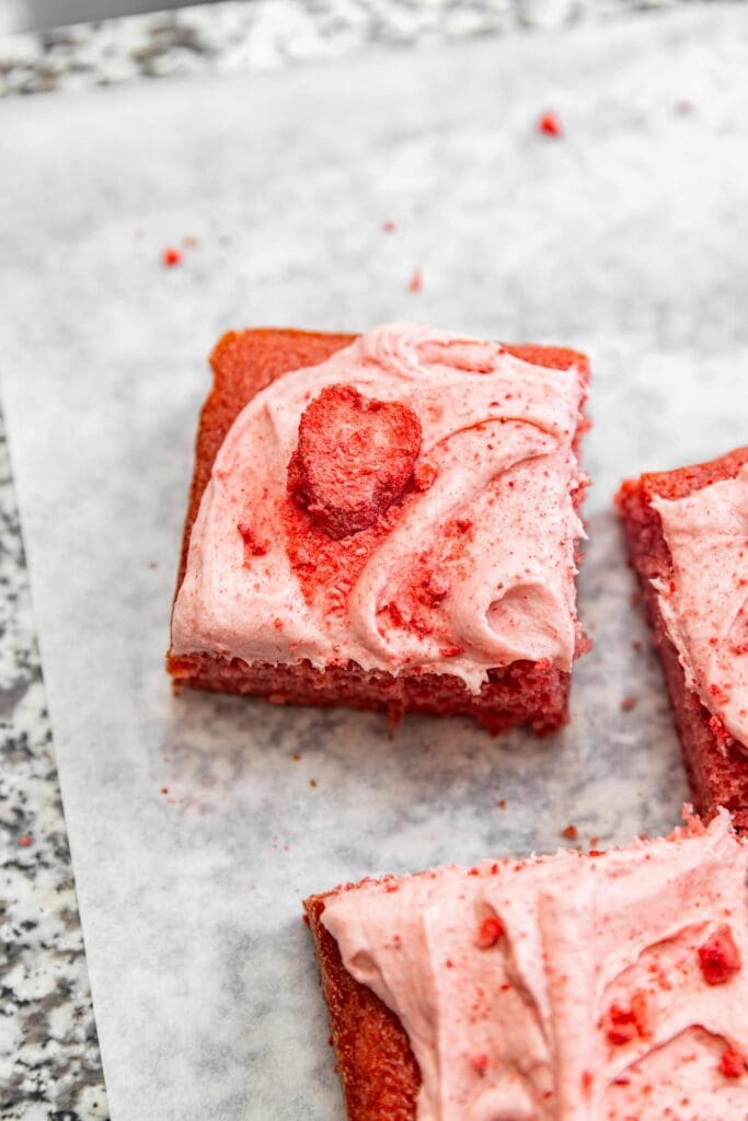 Close up of a slice of strawberry sheet cake with strawberry cream cheese frosting on top and crushed freeze-dried strawberries sprinkled on top.