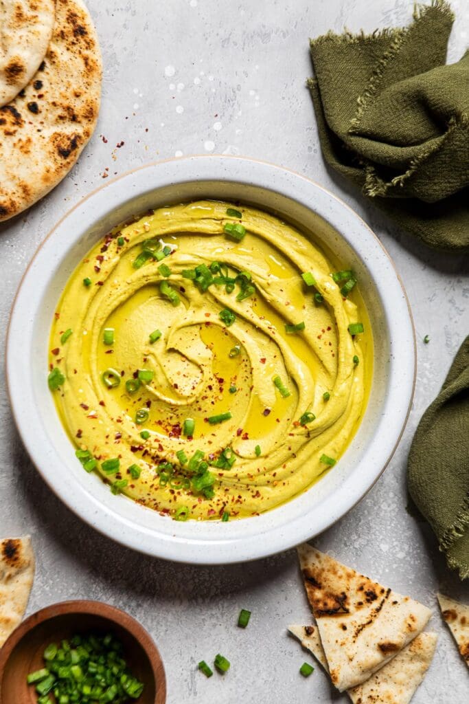 a bowl of curry hummus on a table topped with chopped green onions and chili flakes.