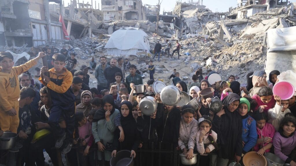 Crowd gathers amid rubble holding containers.