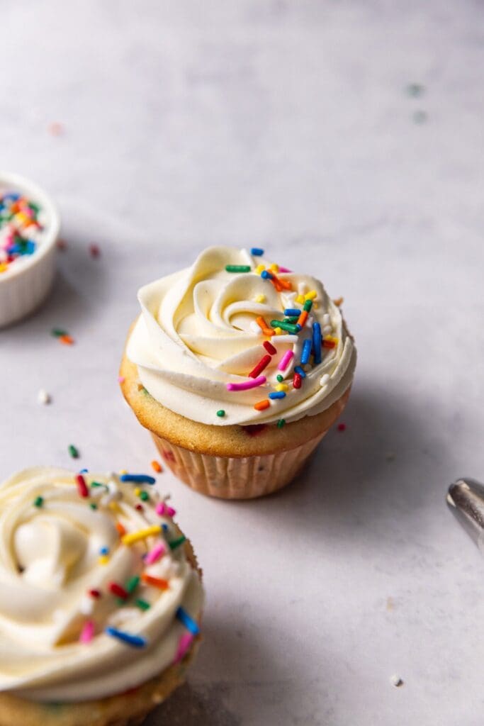 Two funfetti cupcakes with vanilla buttercream frosting and rainbow sprinkles on top sitting next to a small pinch pot of sprinkles and a piping bag filled with frosting.