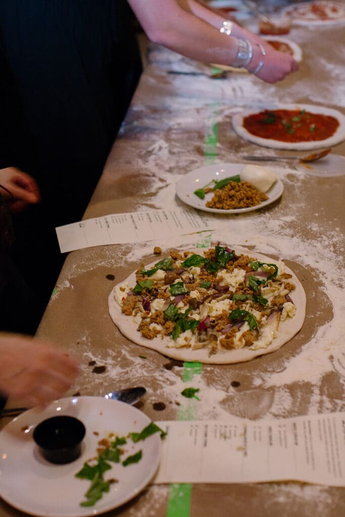 Consuming With Erica Dinner Club: Pizza Making Experience People preparing homemade pizzas with toppings.