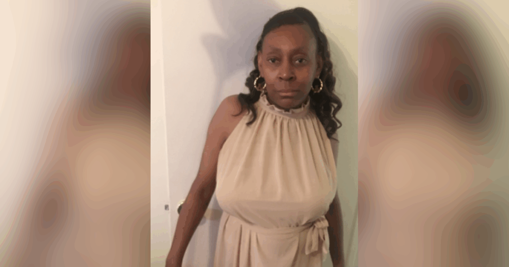 Woman in beige dress standing against wall.