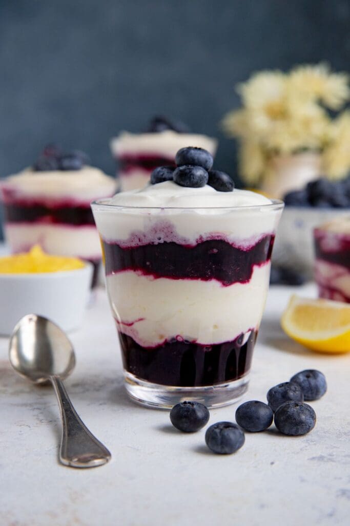 Blueberry lemon parfait in a glass jar with a spoon next to the container.  There are 2 layers of blueberry compote with a layer of lemon cream in between.  The dessert is topped with whipped cream and fresh blueberries.