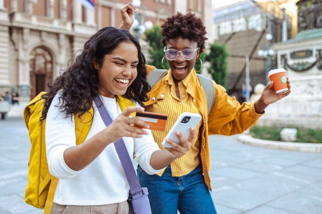Two cheerful people outdoors one showing a credit card and phone while the other holds a coffee cup and gestures excitedly