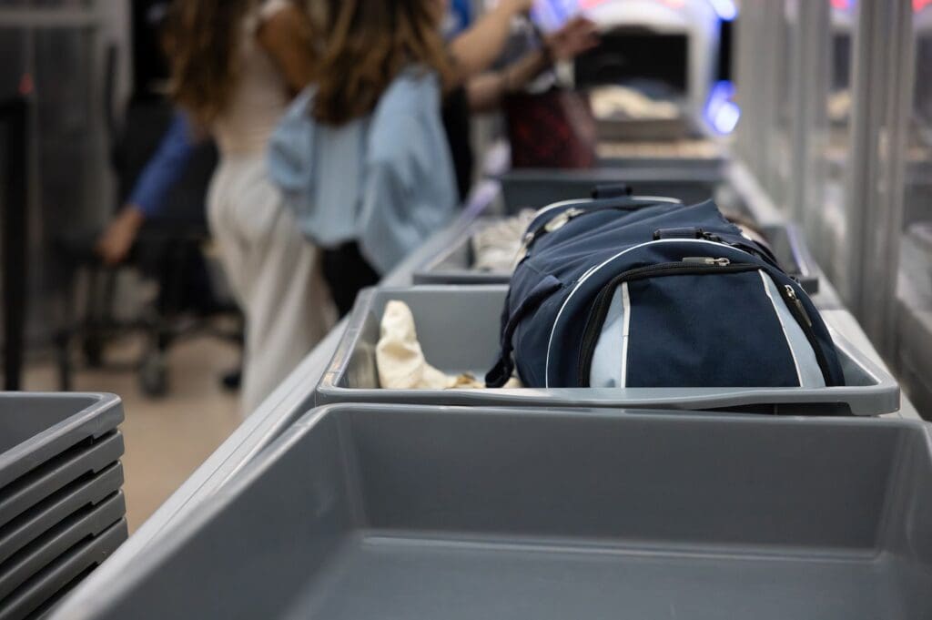 Airport security bins on a conveyor belt with a bag placed inside people visible in the background at a distance