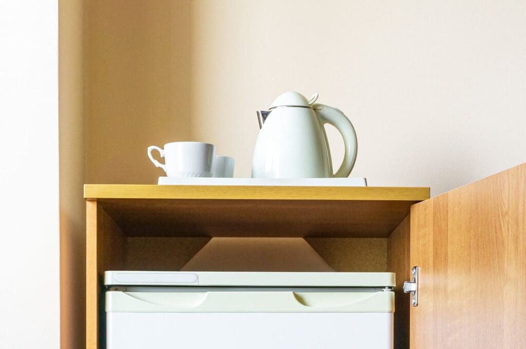 A hotel kettle and cups on a tray placed atop a mini refrigerator in a wooden cabinet