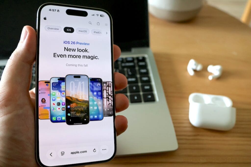 holding iPhone with iOS26 preview on screen. laptop and headphones on desk