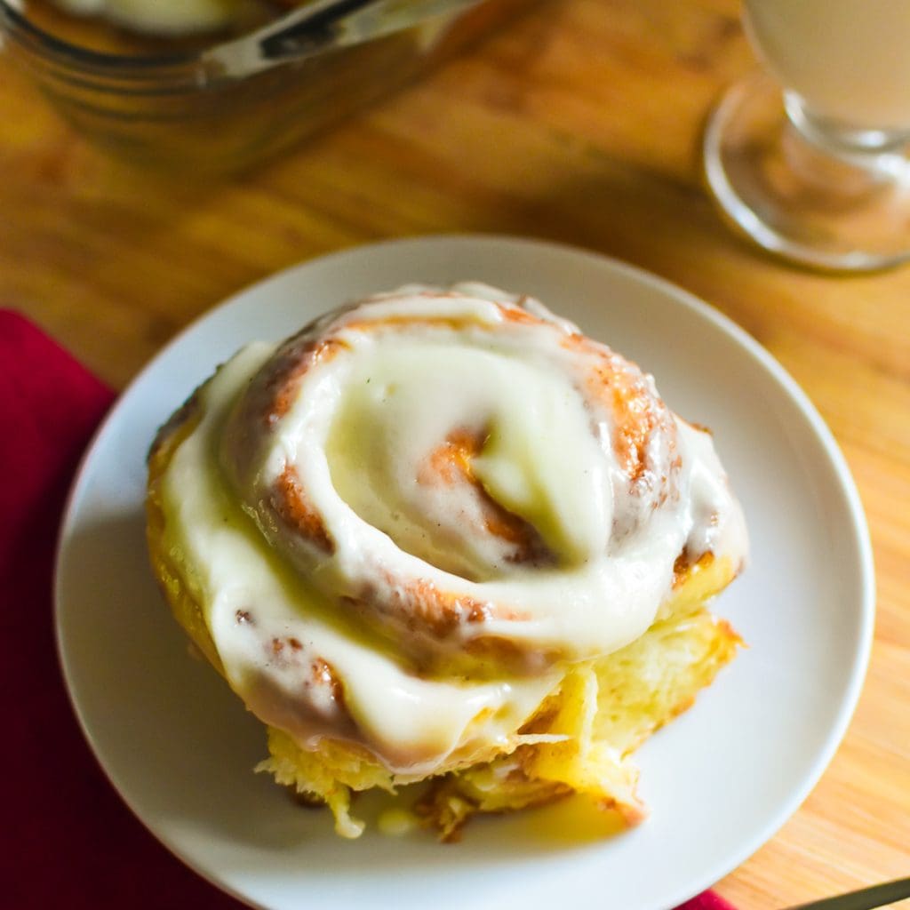 the best soft and fluffy buttermilk cinnamon roll glazed with bourbon cream cheese frosting on a white saucer next to a glass of milk.