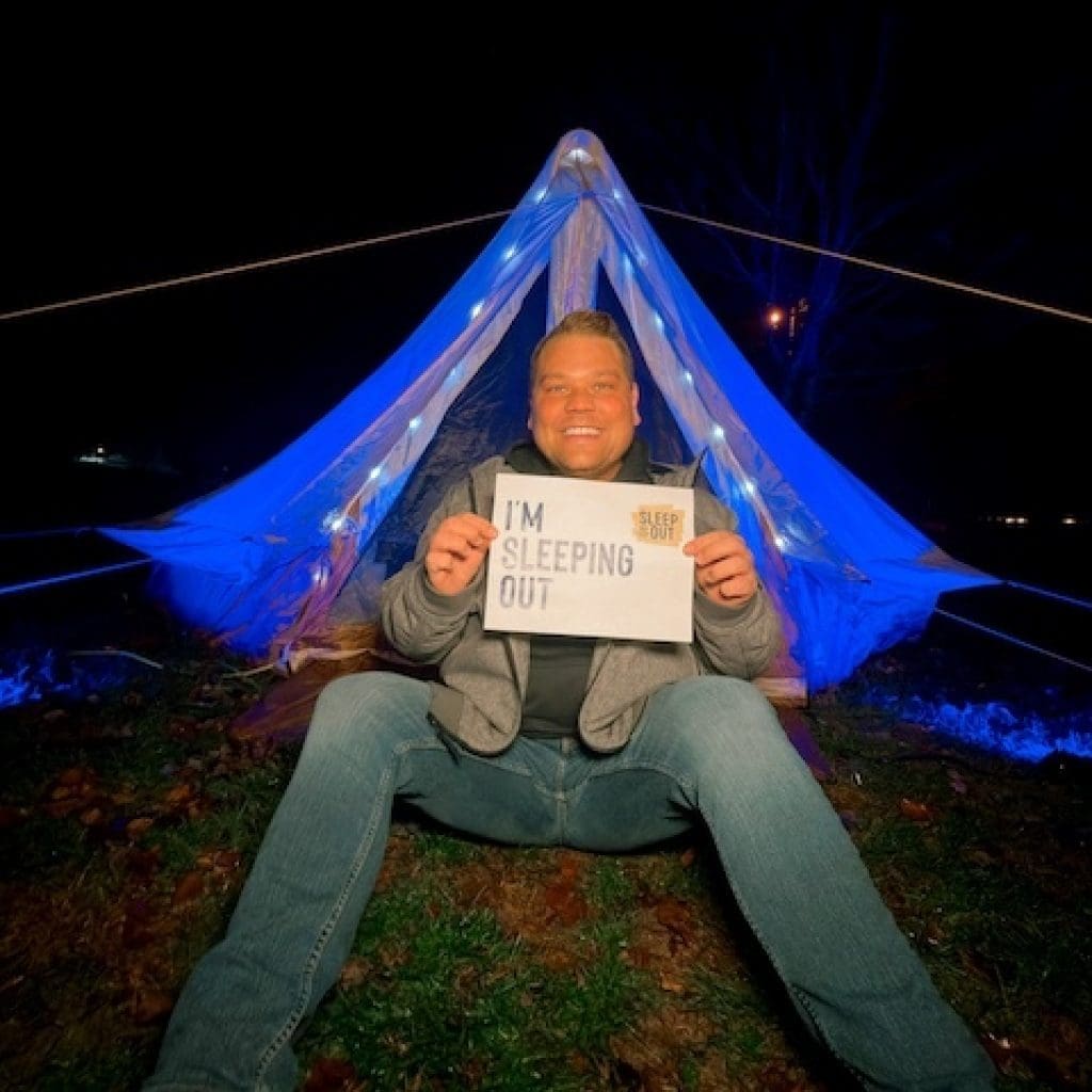 A man, Tyler Mizak, sits smiling in the dark in front of a glowing, blue tent decorated with string lights. He is participating in a Covenant House Sleep Out fundraiser and holds a sign that reads, "I