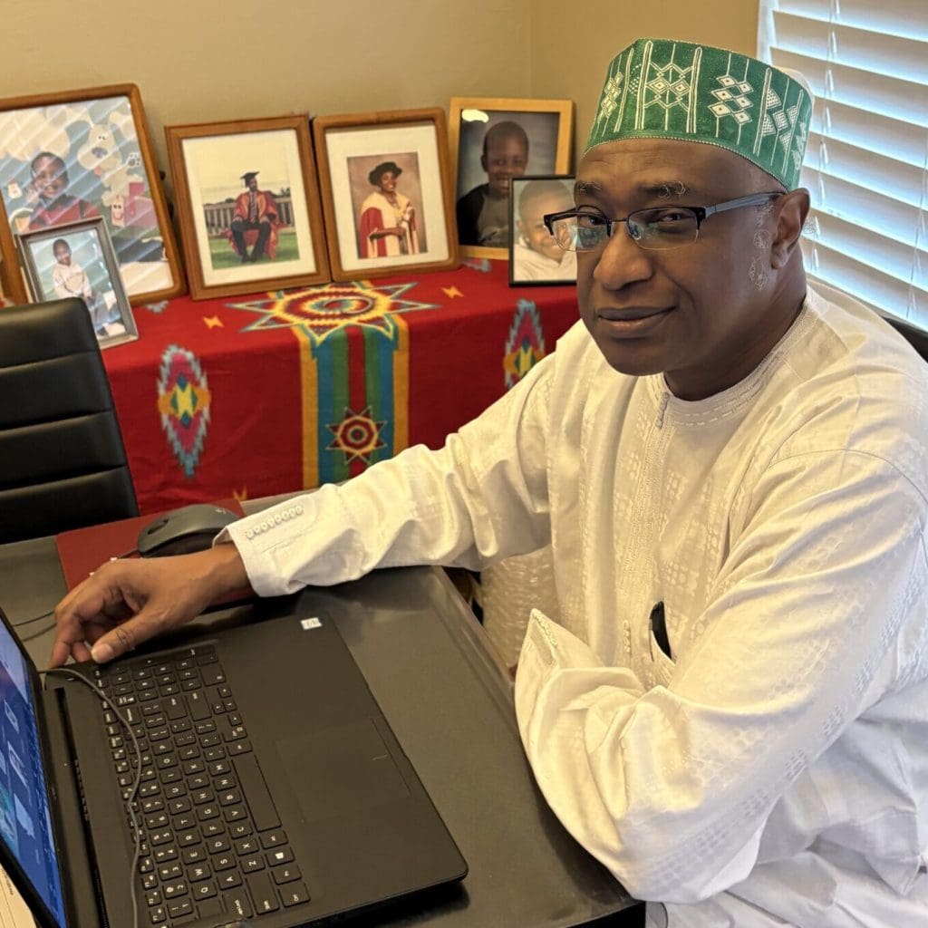 Dr. Mambula sits at his desk wearing traditional Nigerian clothes
