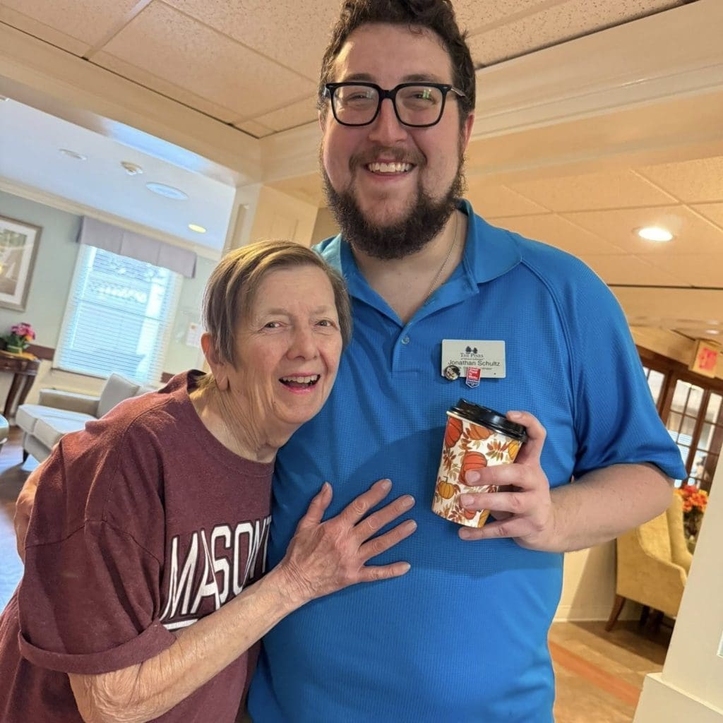 A smiling man with glasses, a beard, and a blue polo shirt standing next to an elderly woman in a maroon