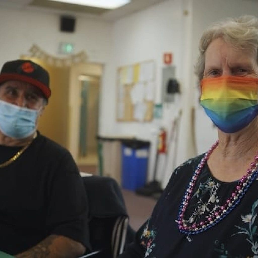 The image shows two individuals sitting together in a communal room in an adult day center. The person on the right, Bonnie, is an older white woman in her 70s or 80s. She is wearing a face mask with a rainbow design and dressed in a black top with a floral pattern. The person on the left, Michael, is wearing a blue surgical mask and a cap, a tee shirt, and gold chain necklace.