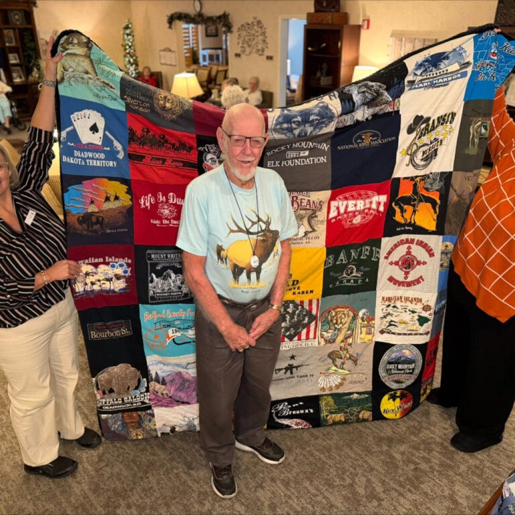 A man and two women showcasing a custom-made t-shirt memory quilt featuring travel and nature-themed graphics.