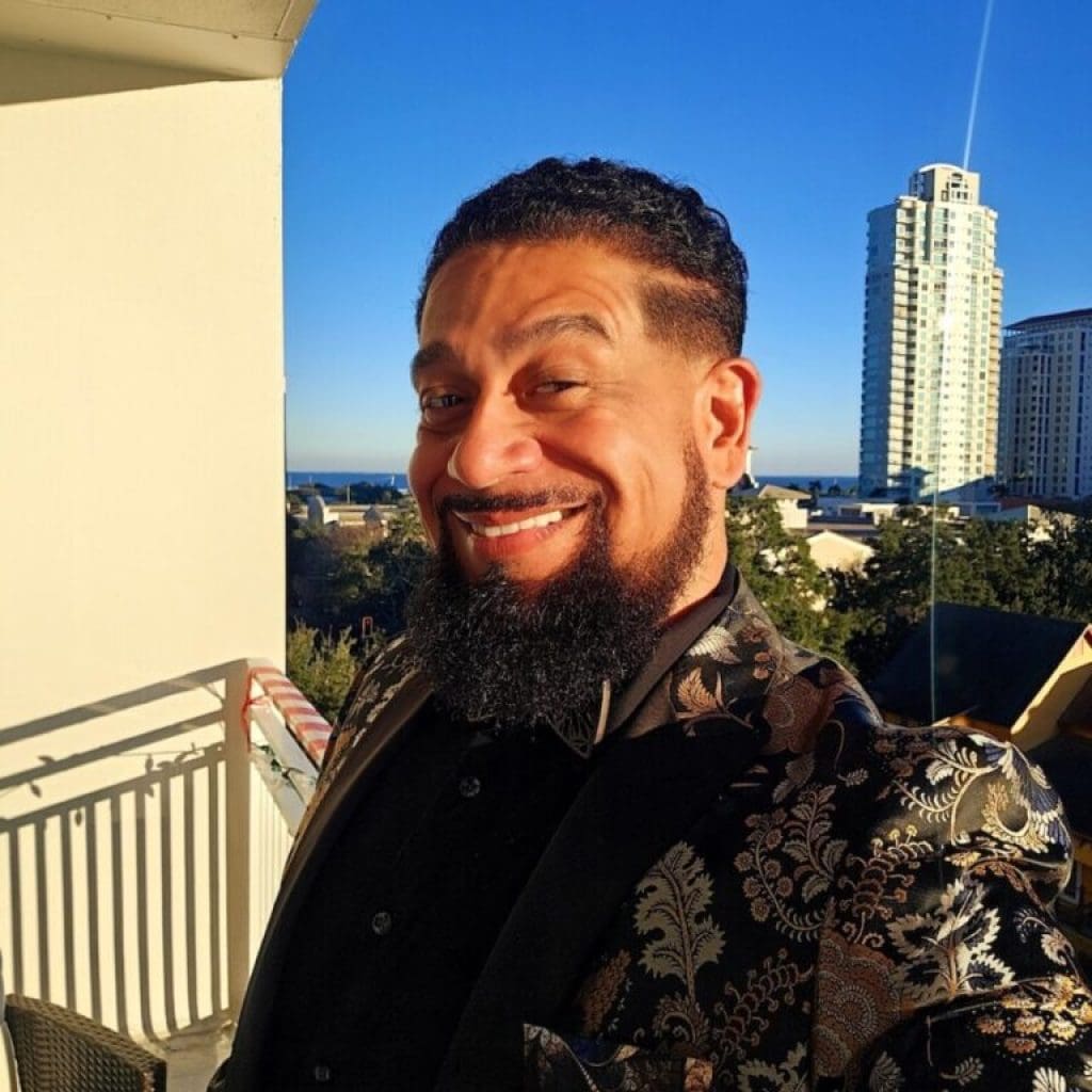 A smiling man with a beard standing on a balcony with a cityscape background.