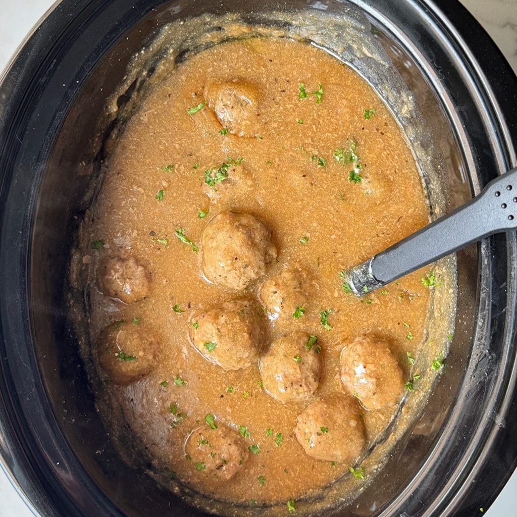 overhead shot of cooked meatballs and gravy in a black slow cooker garnished with fresh parsley