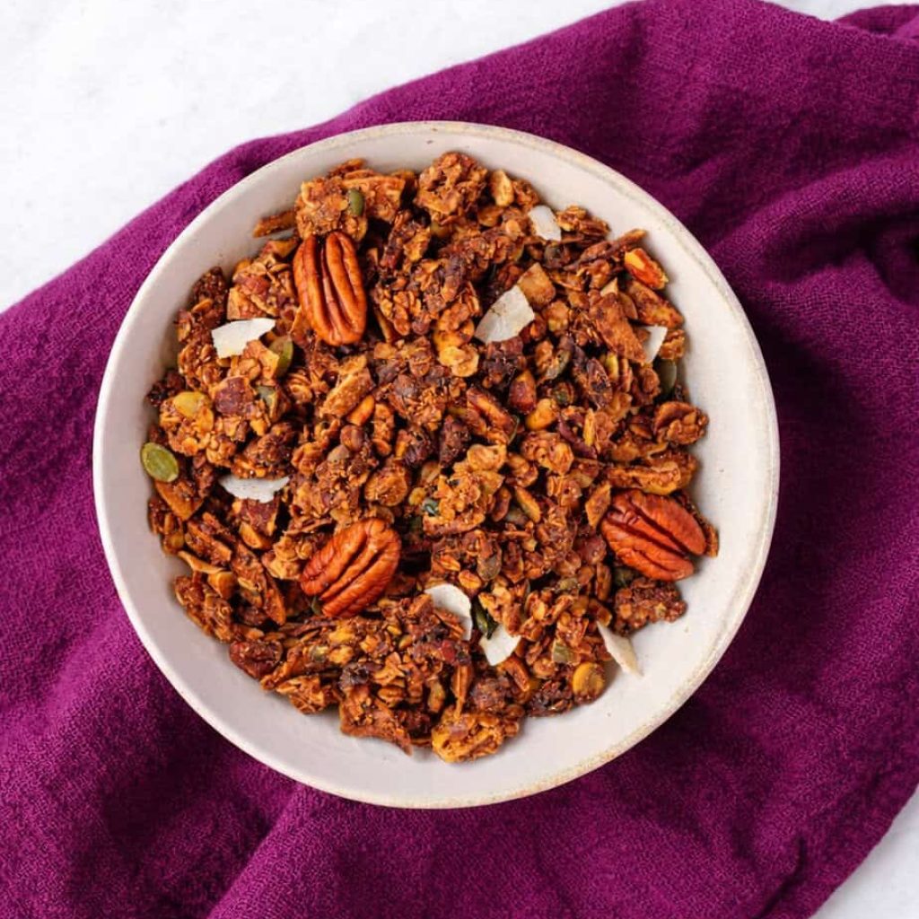 A bowl of granola with pecans and coconut flakes on a purple cloth.