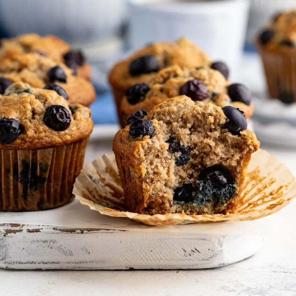 Six blueberry banana muffins on a white board with a bowl of blueberries in the background.  A muffin in the front has a bite taken out of it.