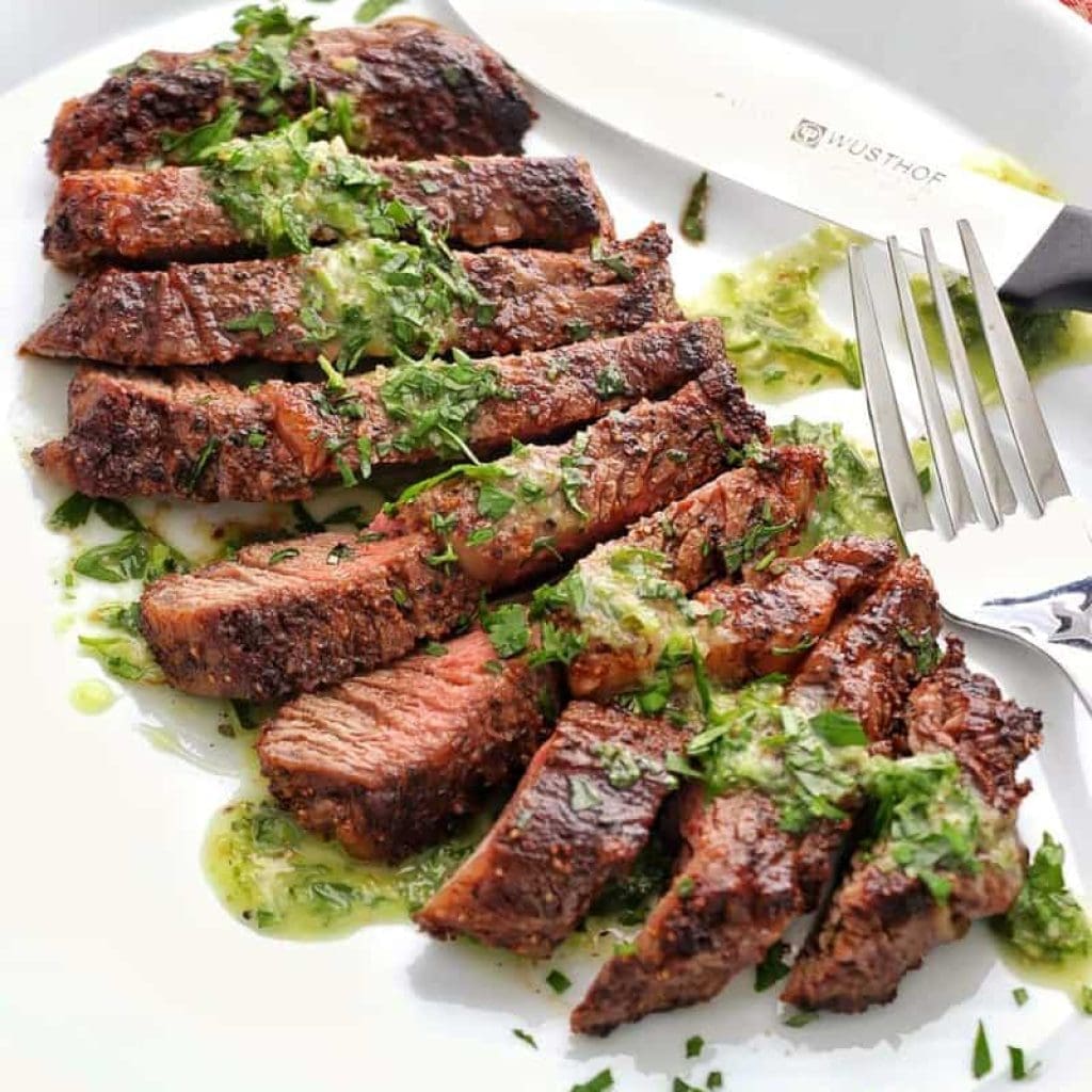 Sliced grilled steak topped with green herb sauce on a white plate.