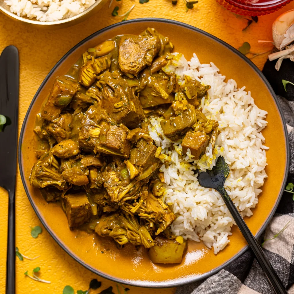 Plant-Based Jamaican Curry Chicken with Jackfruit on a plate with rice and a fork on a yellow table.