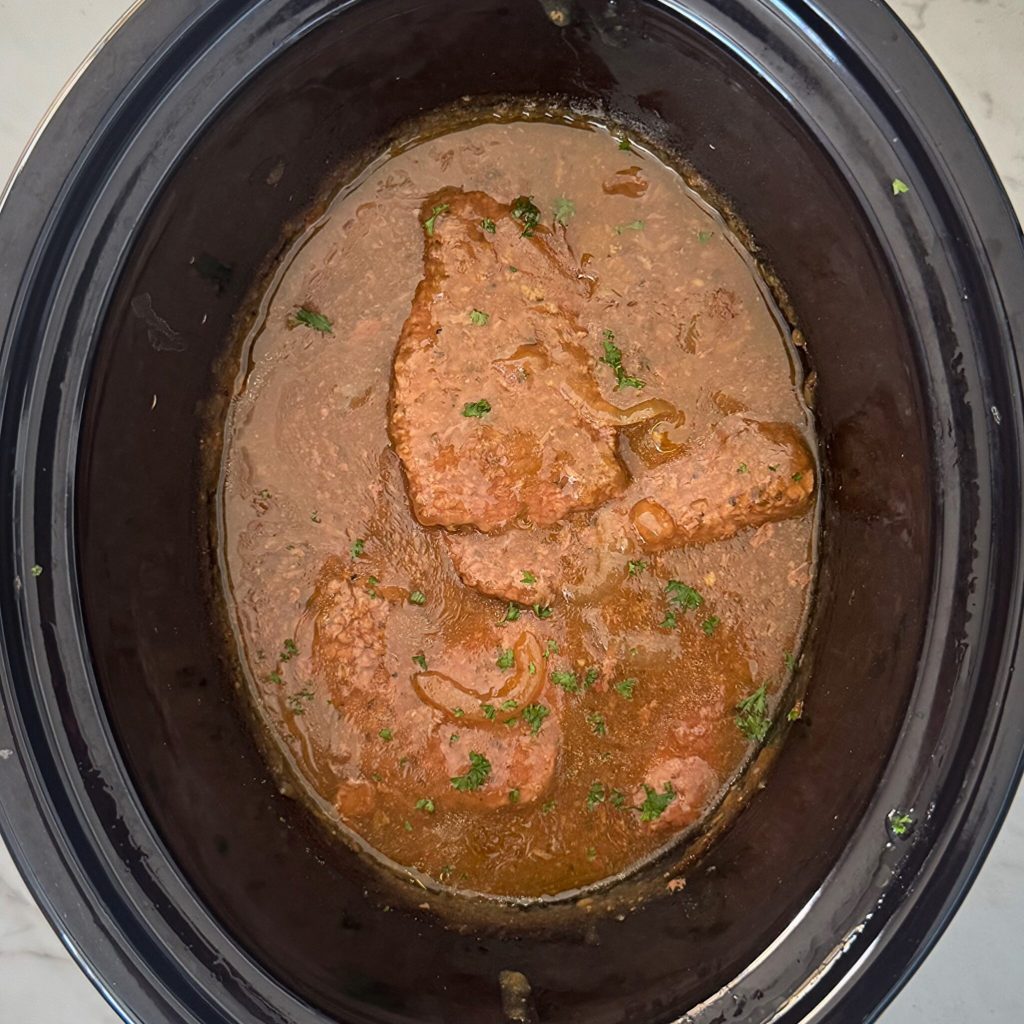 overhead shot of cooked cube steak in a black slow cooker garnished with fresh parsley