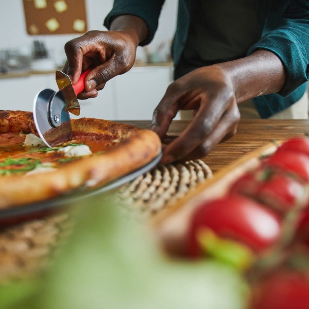 lose-up of African man standing at the table and cutting homemade pizza with knife in the kitchen