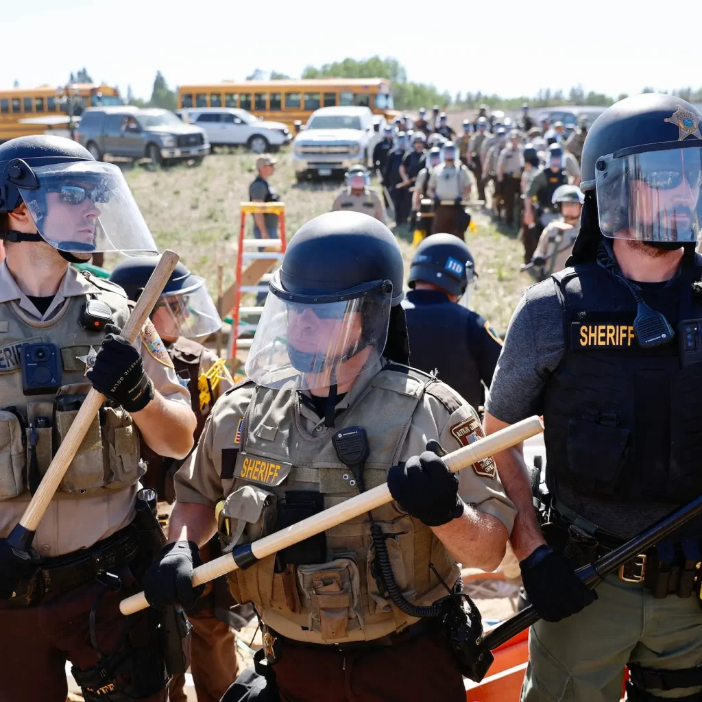 Lines of people in rio gear and sheriff vests and holding bats near yellow school buses in a field