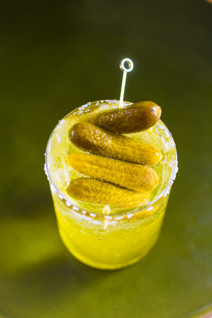 Simple Pickle Spritz Cocktail – Dash of Jazz pickle spritz cocktail in a modern highball glass rimmed with coarse salt and garnished with cocktail pickles.
