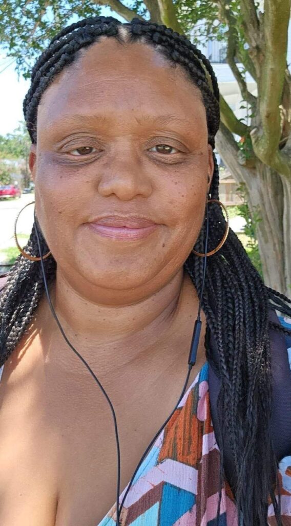 Close-up of a smiling woman with braided hair, gold hoop earrings, colorful top, and earphones outdoors.