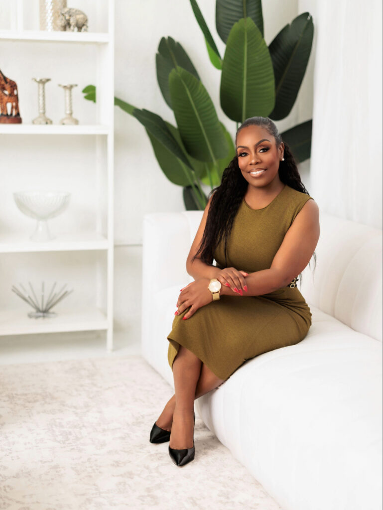 Natasha Blair on Luxury Hospitality, Real Estate Innovation, and Building Exceptional Guest Experiences Through Naki Properties Smiling woman in an olive sleeveless dress sits on a white sofa in a bright living room with large green plants behind her.