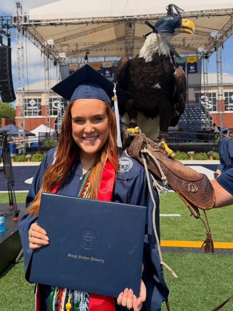 Georgia Southern graduate uses history to leave a legacy Jaylee Kilgo at commencement wearing Georgia Southern branded regalia and holding a diploma cover with Freedom II the eagle behind her shoulder.