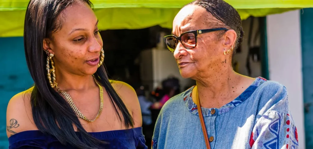 A photo of two Brown-skinned women who may be related, having a conversation outdoors. The younger woman on the left wears a blue off-the-shoulder top, large hoop earrings, and a gold necklace, and has a tattoo on her upper arm. The older woman on the right is wearing glasses and a blue denim shirt.