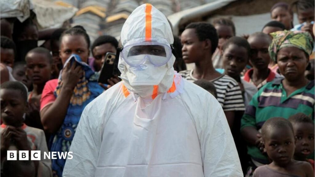UK humanitarian worker monitored for Ebola in London hospital A person in PPE and full head covering is having their goggles adjusted by someone, with blue protective gloves, out-of-focus in the foreground. The green scrubs and hair covering of that person can be seen on the right of the picture.