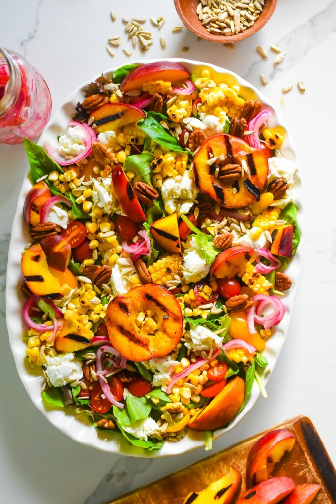 Big Summer Salad with Grilled Peaches and Strawberry Vinaigrette delicious and colorful summer salad with grilled peaches and corn, crunchy nuts and seeds, and goat cheese on a white scalloped platter.