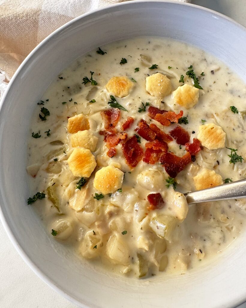 Easy Slow Cooker Clam Chowder close up shot of cooked clam chowder in a white bowl garnished with fresh parsley, bacon, and oyster crackers