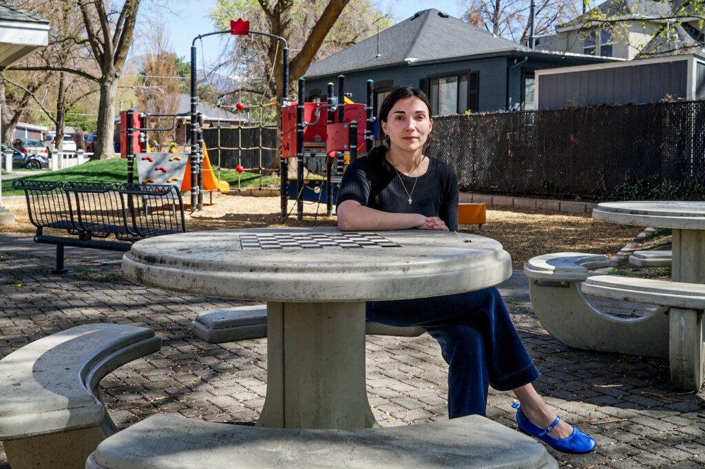 ‘The Child Is Terrified’: Doctors on the Front Lines of a Measles Comeback Speak Out A woman wearing a dark shirt and pants, a bright blue shoes sits at a stone chess table in a park and looks at the camera. A playground is in the background.