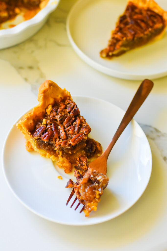 slice of the best old fashioned gooey pecan pie made with corn syrup, vanilla, and bourbon, served on a white saucer with a copper fork.