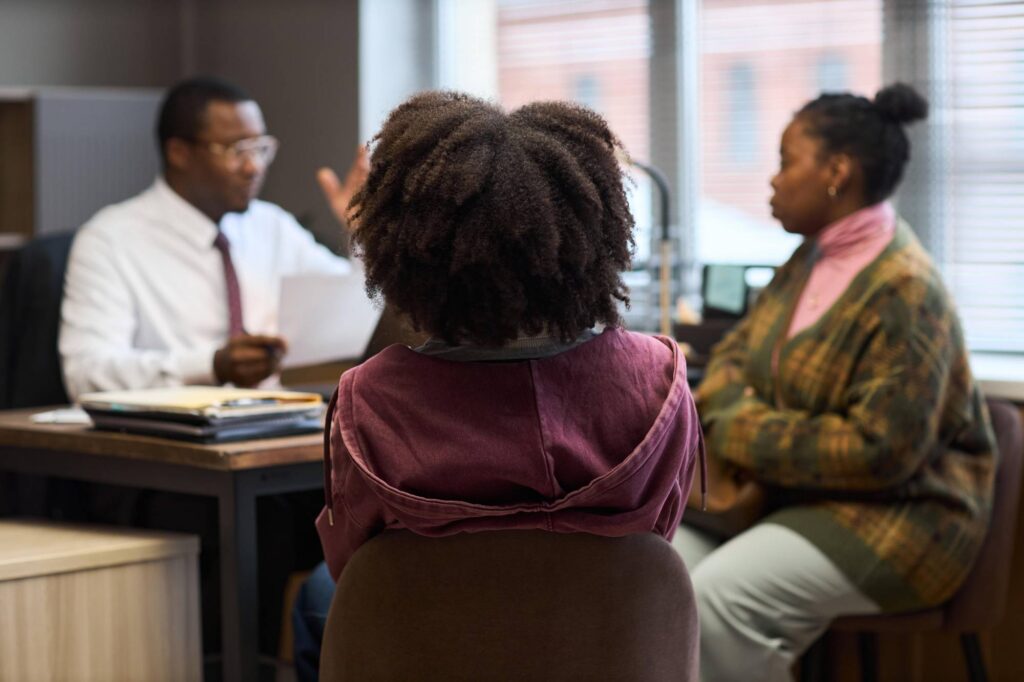 Black man appearing middle aged sitting at desk talking to Black woman and Black child in office setting, principal discussing school matters with parent and student.