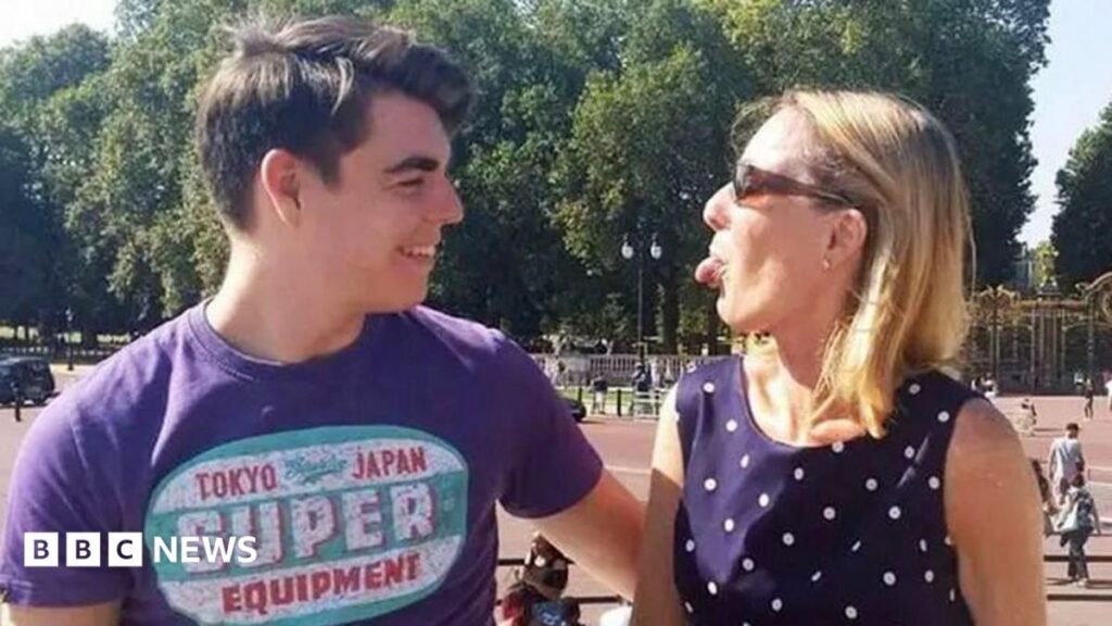 NHS staff investigated over access to dead teenager’s medical records Oliver McGowan, wearing a purple t-shirt with a logo, is standing outside Buckingham Palace. He is starring at his mother Paula McGowan who is standing next to him and wearing a dark blue polka dot dress and sunglasses. She is playfully sticking her tongue out at her son and he is laughing.