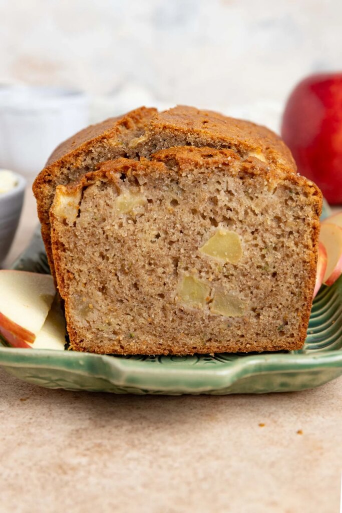 A close-up of a sliced loaf of apple zucchini bread on a white plate, showing a moist texture with apple chunks and small bits of zucchini visible throughout.