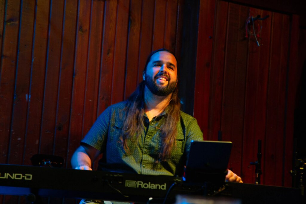 Nonprofits Are Helping Musicians Pay for Insurance in Austin, Texas, and Beyond A man with a short beard and long brown hair in a button down green shirt smiles as he plays a keyboard in a music venue.