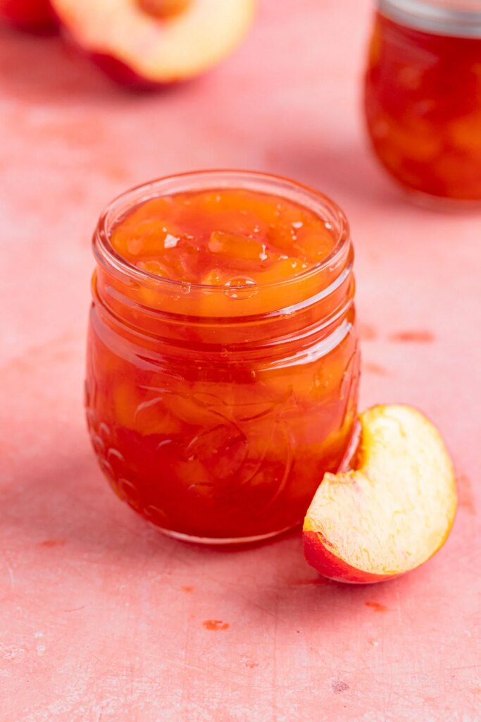 Small Batch Peach Jam (No Pectin, No Canning) An 8-ounce open jar of homemade peach jam with a small silver spoon next to the jar. There is a another jar of peach jam in the background. There are fresh peaches next to the open jar and in the background.