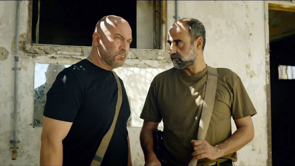 "Two serious men with shoulder bags stand in front of a weathered, sunlit building, partially shaded by window light inside a worn, possibly abandoned environment."